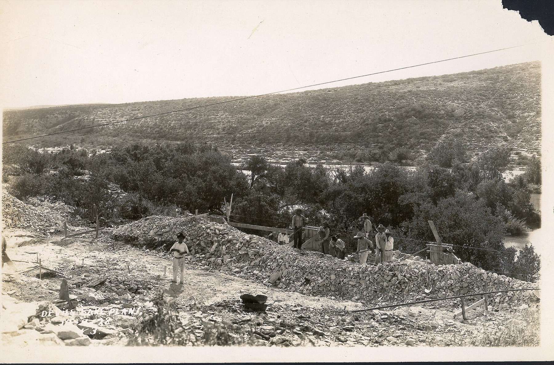 Black and white photo of a man standing in front of and several men digging rock on top of a large berm of rock parallel to a river